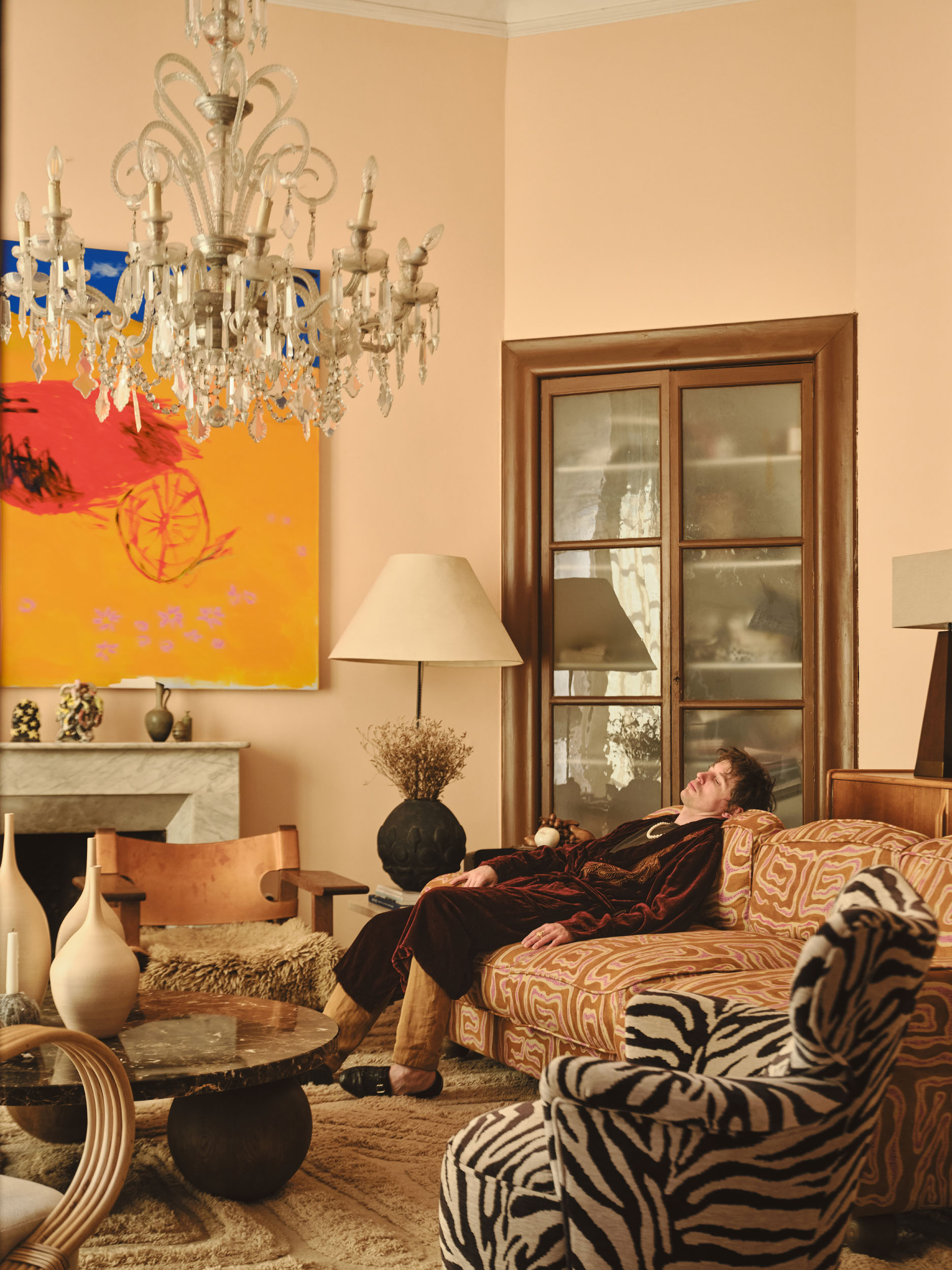 Robert Mollard photographed by Andrea Papini at Arcadian Landscapes, Dusty Deco, Palma de Mallorca, 2026. Curated by The Art Bystander. The artist reclines on a patterned sofa beneath a crystal chandelier, wearing a dark velvet robe. Behind him, his large-scale painting Massacre of the High-Score hangs above a marble fireplace.