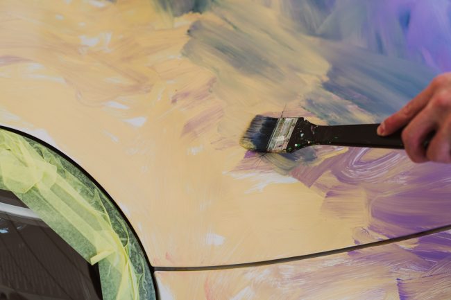 Close-up of a wide brush dragging blue paint across the pale yellow lacquer of the car hood
