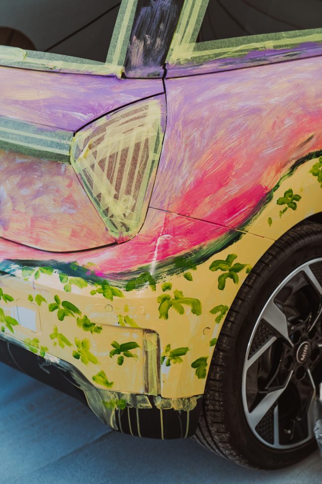 Rear detail of the MINI Art Car with masking tape still on, showing a pink sky and green palm trees painted over a yellow field