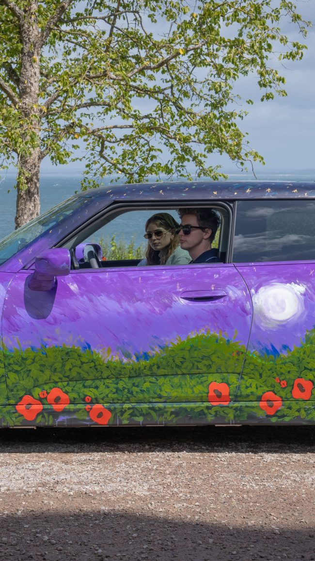 Robert and Madelen Möllard seen through the side window driving the MINI Art Car, a painted moon and poppies on the purple door