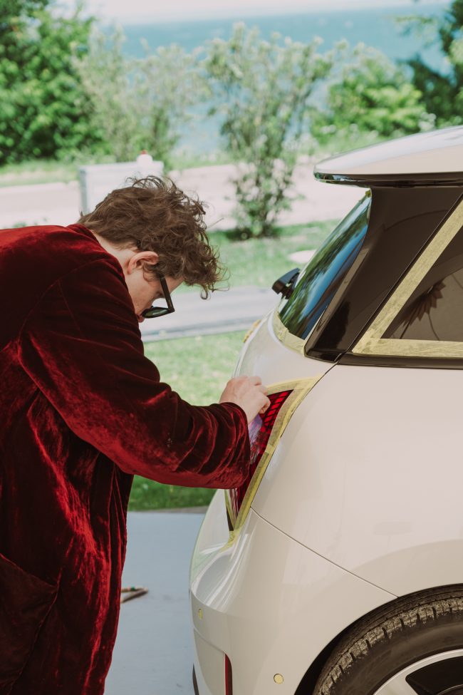 Robert Möllard in a dark red velvet robe masking the tail light of a white MINI Cooper before painting