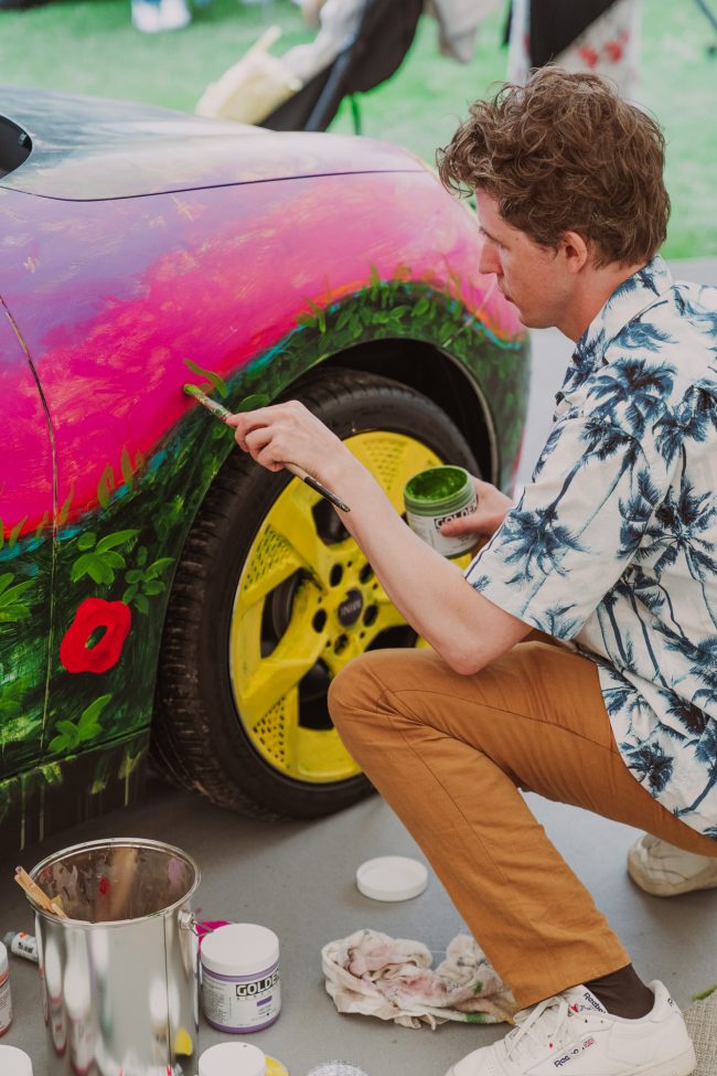 Robert Möllard crouching by the wheel arch of the MINI Art Car, painting green foliage over a pink sky during the live painting