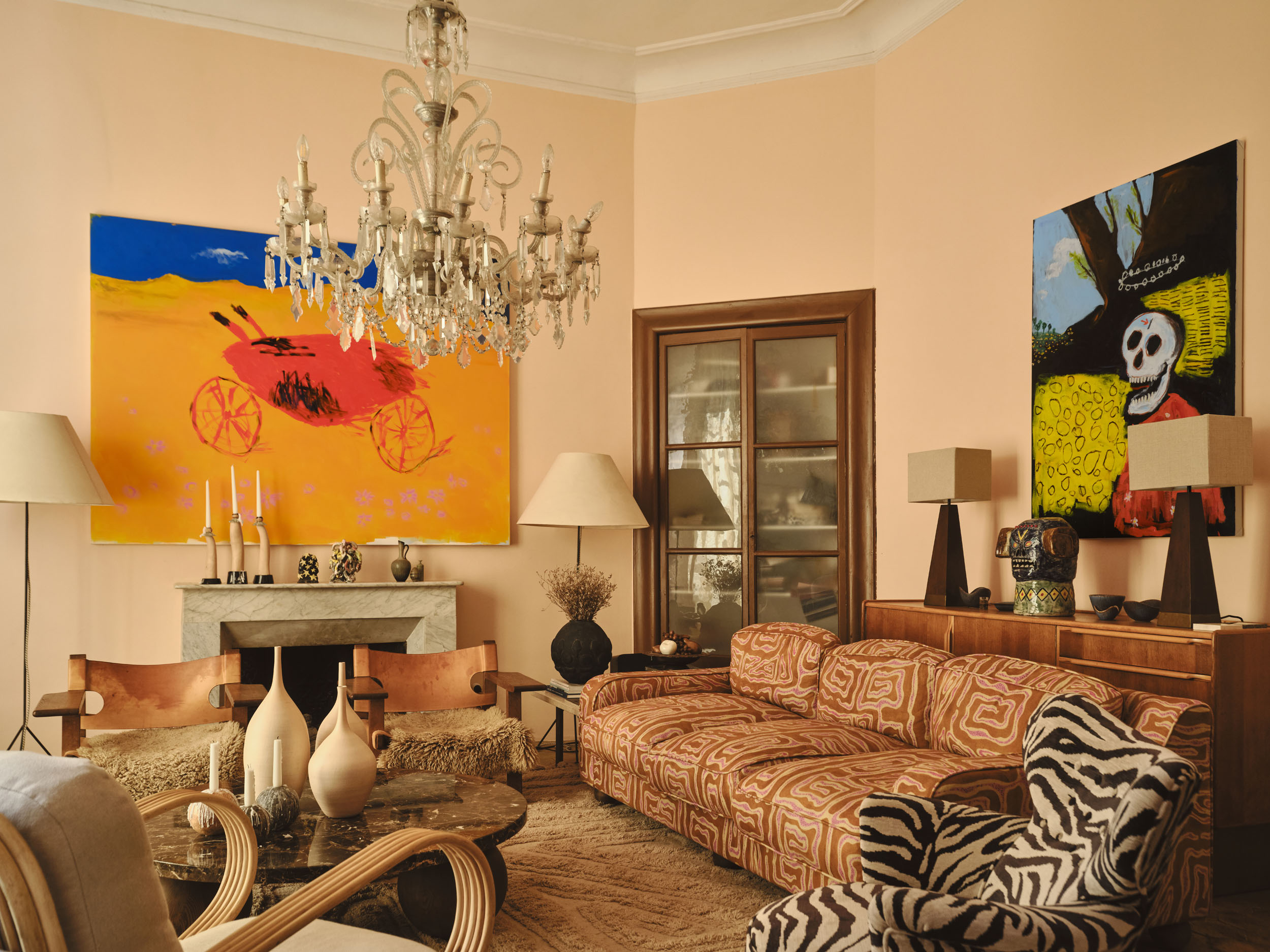 Robert Möllard, installation view from Arcadian Landscapes, Dusty Deco Showroom, Palma de Mallorca, 2026. A peach-walled salon with a marble fireplace and a crystal chandelier. Massacre of the High-Score hangs above the fireplace, a red arcade motorcycle crossing a golden yellow ground beneath a deep blue sky. On the adjacent wall hangs Et in Arcade ego, a haloed skull in a red coat painted with white daisies, flanked by yellow fields of coin shapes. Photography by Andrea Papini.